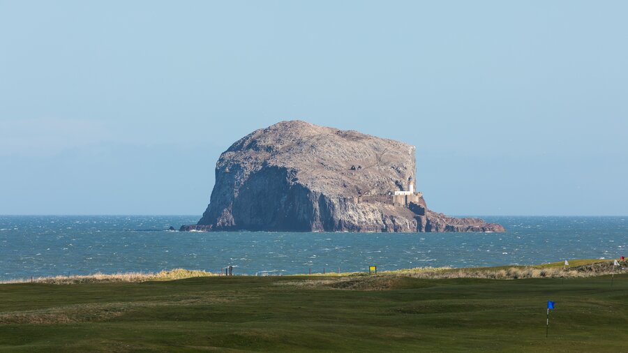 Bass Rock viewed from North Berwick - A large rock looms from the water with a grassy shoreline in the foreground. (© Visit Scotland)